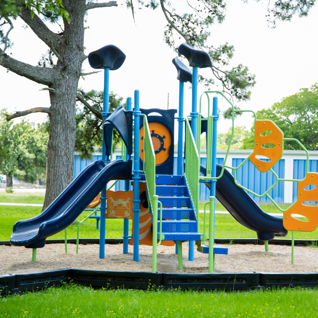 A colorful playground structure with slides and climbing features set in a grassy area under trees.