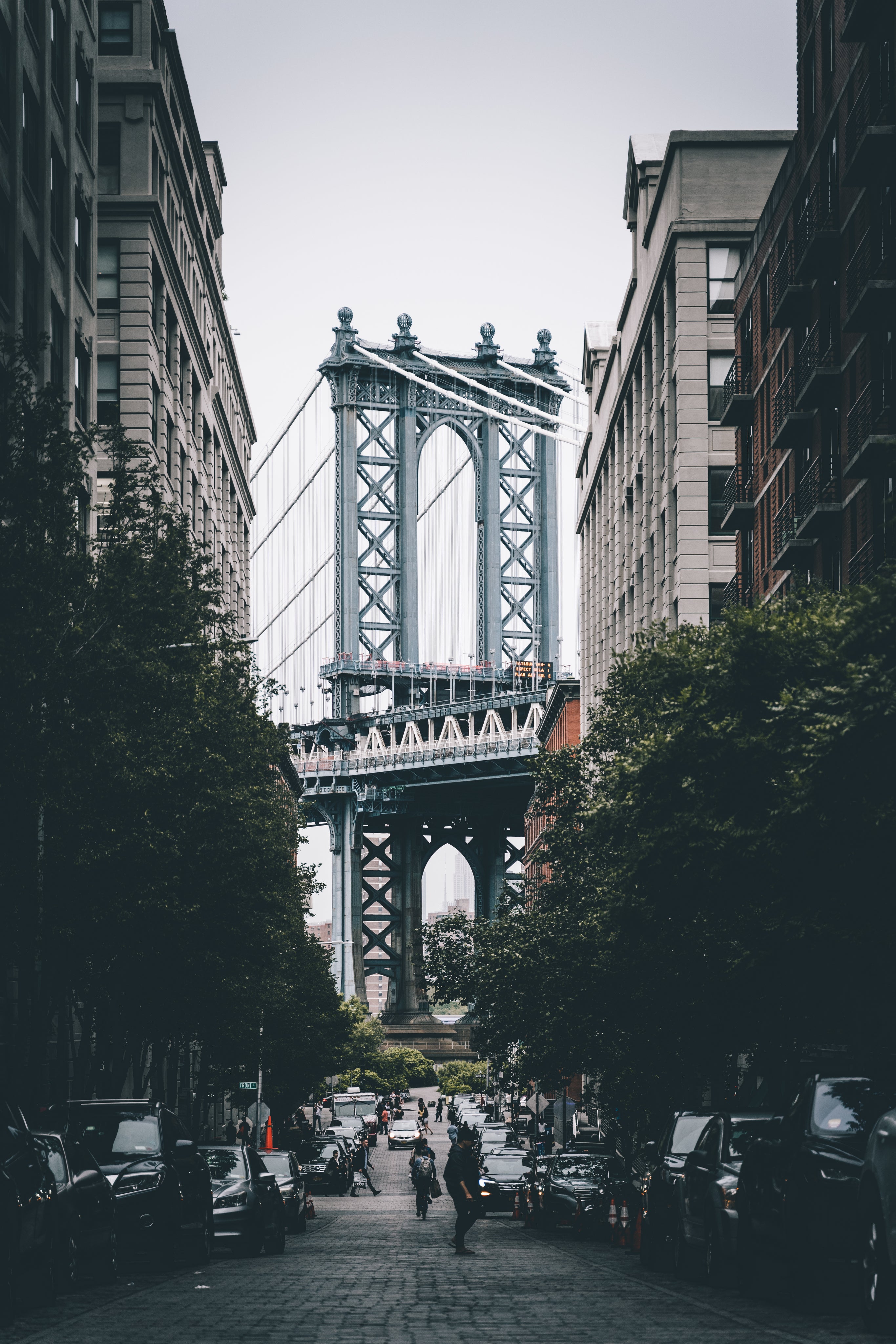 View of the Manhattan Bridge framed by buildings, with a street and parked cars in the foreground.