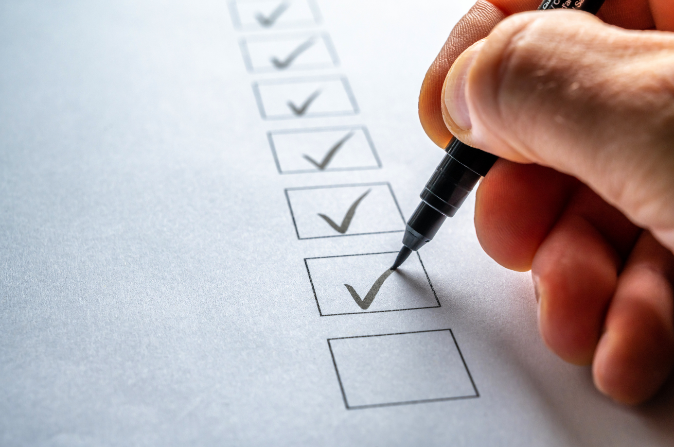 Hand with pen marking checkboxes on a paper checklist. Eight boxes, four checked. Image suggests organization, completion, and productivity.