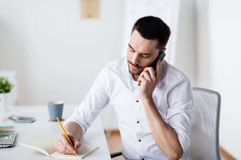 A man in a white shirt sitting at a desk, talking on the phone while writing in a notebook.