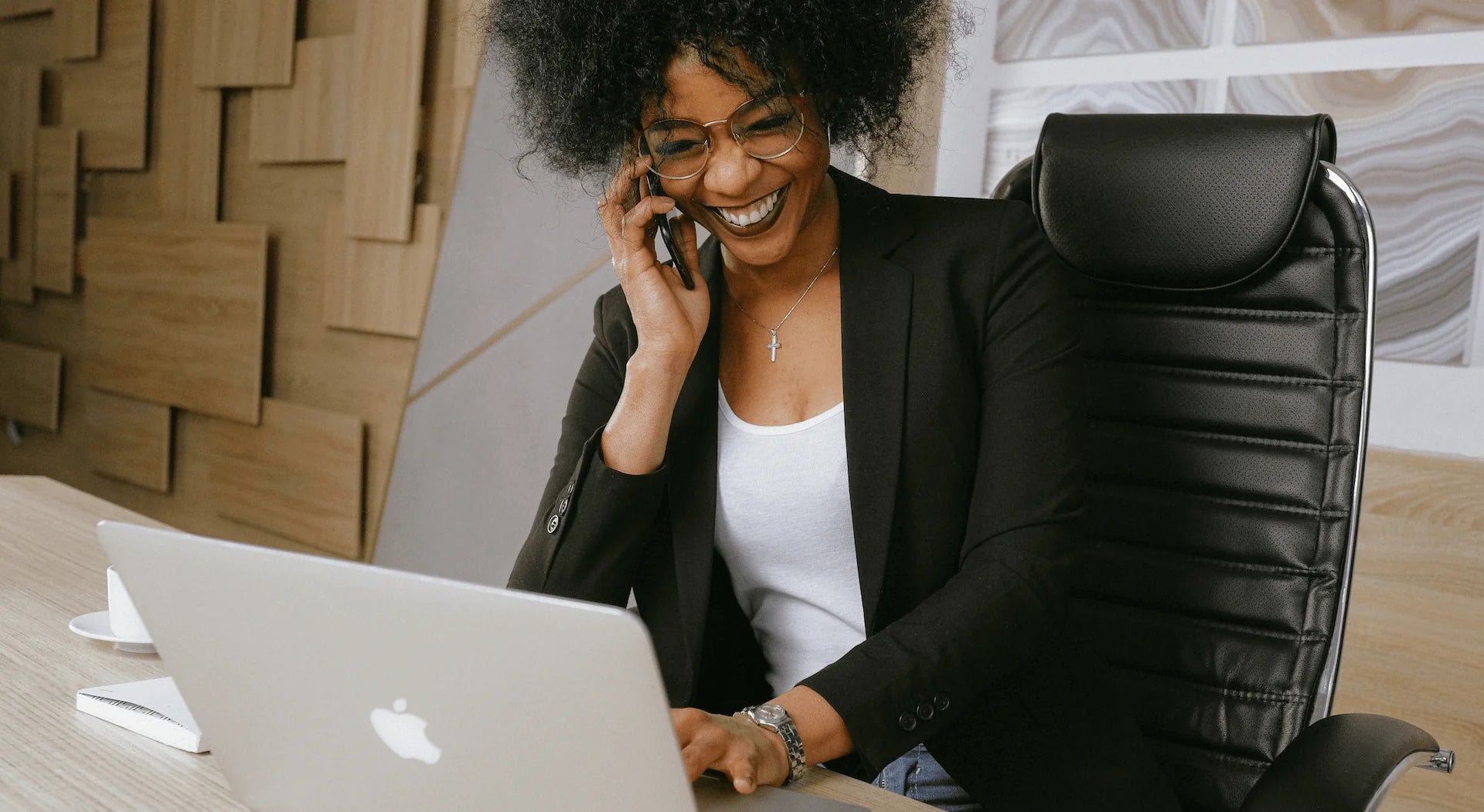 Woman in a black blazer smiles while talking on the phone and working on a laptop at a desk.